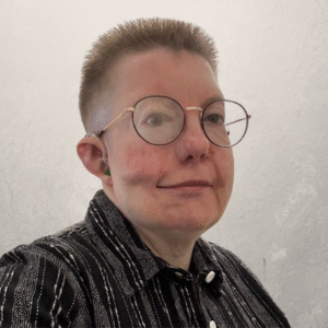 White person with a skin fade style hair cut wearing glasses, a hearing aid with green earmold and a black button up shirt with white vertical stripes facing to the right but with head turned forwards towards the camera